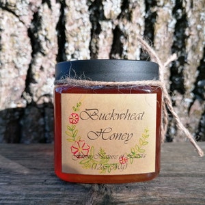 Könnte beinhalten: Ein durchsichtiges Glas mit bernsteinfarbenem Buchweizenhonig, einem schwarzen Deckel und einem beigen Papieretikett. Das Etikett zeigt den Text Buckwheat Honey, From Nature of Latvia und 12oz 340g, verziert mit einer Blumenillustration und Bindfaden.