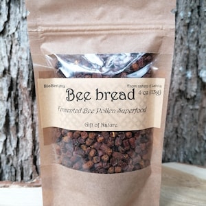 Puede incluir: Una bolsa de papel marrón que contiene pan de abeja de color marrón oscuro, un superalimento de polen de abeja fermentado. La etiqueta dice BioBeeLatvia, Bee bread, 4 oz (113g), Fermented Bee Pollen Superfood y Gift of Nature. La bolsa tiene una ventana.