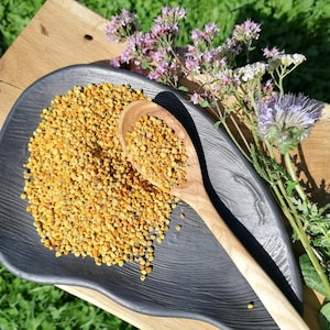 May include: A pile of golden yellow bee pollen granules rests on a dark grey, organic-shaped ceramic dish. A light-coloured wooden spoon filled with pollen sits atop the pile, accompanied by a sprig of purple and white wildflowers nearby.
