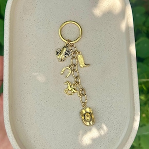 Könnte beinhalten: Goldfarbener Schlüsselanhänger mit mehreren Anhängern, darunter ein Kaktus, ein Hufeisen, ein Cowboy-Stiefel, ein Pferd und ein Cowboyhut. Die Anhänger sind durch eine Kette verbunden und an einem runden Schlüsselring befestigt. Der Schlüsselanhänger wird auf einem hellen, ovalen Tablett präsentiert.