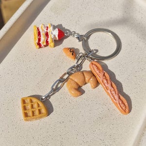 Könnte beinhalten: Ein silberner Schlüsselanhänger mit Miniatur-Essensanhängern. Die Anhänger umfassen eine Waffel, ein Croissant, ein Baguette und ein Stück Kuchen. Das Kuchenstück ist gelb mit roter und weißer Glasur.