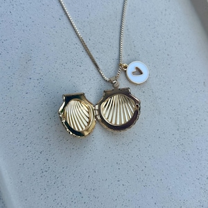 Puede incluir: Collar dorado con un colgante en forma de concha, abierto para revelar el interior. Un pequeño dije blanco con un corazón recortado cuelga cerca. La cadena del collar es delicada. Fondo claro y moteado.