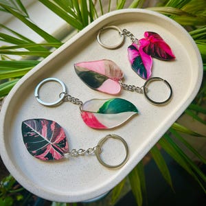 May include: Keychains featuring leaf designs in shades of pink, green, and black. Each keychain has a silver-colored ring and chain. The keychains are arranged on a white, oval-shaped tray, with green foliage in the background.