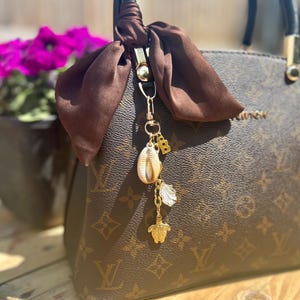 Puede incluir: Un bolso marrón con un charm decorativo. El charm presenta una concha, una tortuga dorada, una pluma blanca y un colgante con la letra "B" dorada. Una bufanda de satén marrón está atada al asa.
