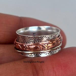 May include: A silver and copper spinner ring with intricate floral designs. The ring is made of three bands that spin freely.