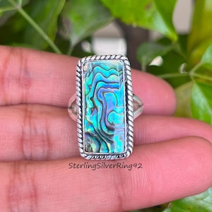 May include: A silver ring featuring a rectangular abalone shell inlay. The shell displays a green, blue, and purple iridescent pattern. SterlingSilverRing92 is printed below the ring.