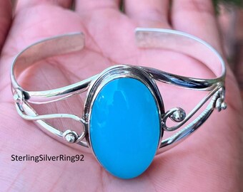 May include: A silver bangle bracelet with a light blue oval gemstone. The bracelet has a delicate, intricate design with a curved shape.