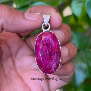 May include: Oval-shaped pendant with a large, faceted ruby gemstone in a silver setting. The ruby is a vibrant shade of pink. The pendant has a loop at the top for a chain. The text "SterlingSilverRing92" is at the bottom.