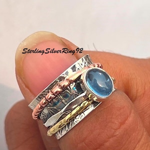 Puede incluir: Un anillo giratorio de plata con una piedra preciosa azul, acentos de cobre y oro. SterlingSilverRing92 está impreso en el anillo.