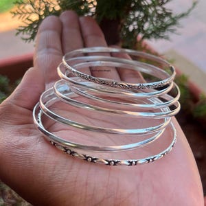 May include: A set of six silver bangles with different designs. The bangles are stacked on top of each other and have a shiny finish.