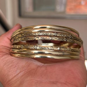 May include: A selection of gold bangles, some with floral patterns, are held in a hand. The bangles have varying widths and designs, with some smooth and others with detailed embossed patterns. The text "SterlingSilverRing92" is visible.