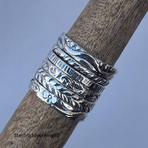 Puede incluir: Primer plano de anillos de plata con diversos diseños grabados, incluyendo patrones florales y geométricos. Los anillos están apilados en una clavija de madera, mostrando los detalles y texturas de la joyería.