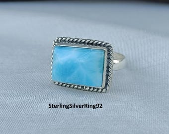 May include: A silver ring featuring a rectangular, pale blue gemstone. The gemstone is set within a silver frame with a twisted rope pattern. The ring rests on a light grey surface. Text on the image: SterlingSilverRing92.