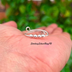 May include: A silver ring featuring three small, spherical beads and a looped design. The ring is held in a hand, with the text "Sterling Silver Ring 92" visible below. The background is blurred green.