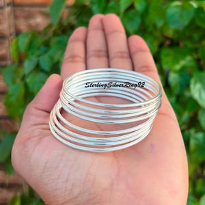 Può includere: Un set di otto braccialetti d'argento impilati uno sopra l'altro. I braccialetti sono lisci e lucidi, e sono tutti della stessa dimensione. Il testo "Sterling Silver Ring 92" è visibile sul braccialetto superiore.