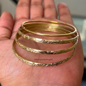 Könnte beinhalten: Mehrere goldfarbene Armreifen, einige mit floralen Mustern, sind übereinander gestapelt. Die Armreifen werden auf der Hand einer Person präsentiert. Der Text "SterlingSilverRing92" ist im Bild zu sehen.