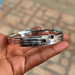 Könnte beinhalten: Ein silbernes Armreif-Armband mit gehämmerter Textur und zwei roten Edelstein-Akzenten. Das Armband hat einen Herz-Anhänger.