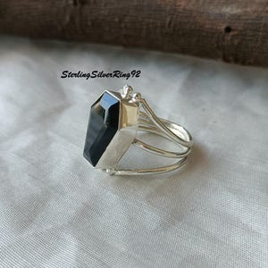 May include: A silver ring with a black gemstone in the shape of a coffin. The ring has a triple band design.