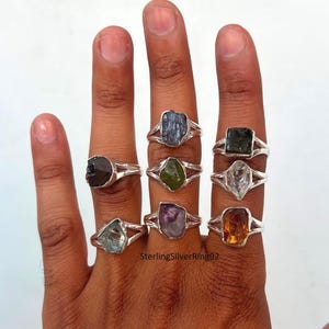 May include: Eight sterling silver rings with different colored gemstones. The rings are on a person's hand. The gemstones are aquamarine, amethyst, citrine, clear quartz, green, black, and blue.