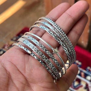 Könnte beinhalten: Ein Set aus sieben silbernen Armreifen mit verschiedenen gravierten Designs, darunter Blumenmuster, ein Stern und eine Wellenlinie. Die Armreifen sind übereinander auf einer Hand gestapelt.