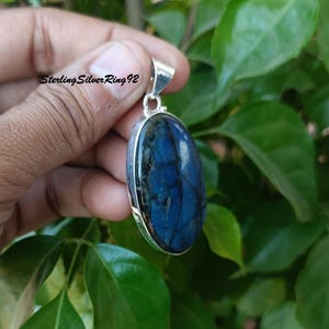 May include: A silver pendant with a large oval-shaped blue labradorite gemstone. The stone has a dark blue color with iridescent flashes of green and gold.