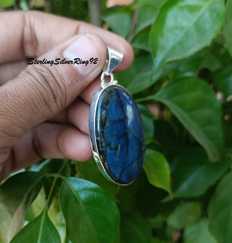 May include: A silver pendant with a large oval-shaped blue labradorite gemstone. The stone has a dark blue color with iridescent flashes of green and gold.