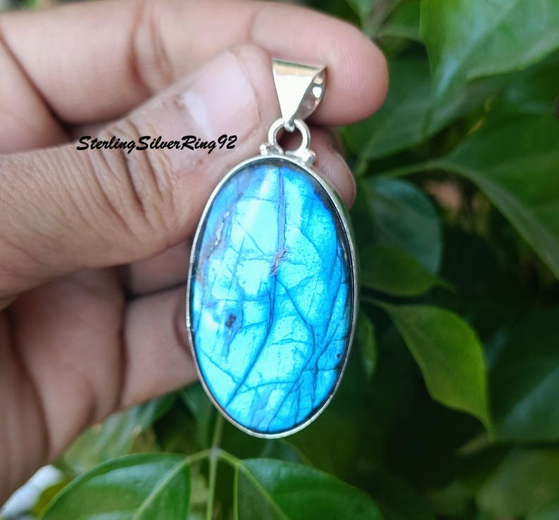 May include: A silver pendant with a large oval-shaped blue labradorite gemstone. The stone has a shimmering iridescent effect.
