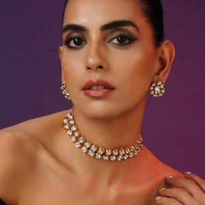 Puede incluir: Una mujer lleva un collar dorado y pendientes a juego. La joyería presenta múltiples piedras redondas y transparentes engastadas en un metal dorado. El fondo es un degradado de morado y rosa.