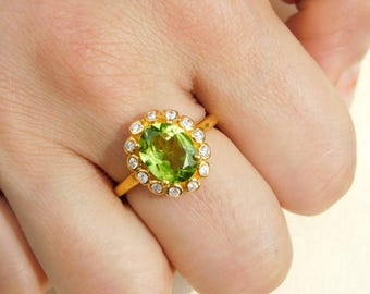 May include: A gold ring featuring a large, oval-cut, light green gemstone, surrounded by a halo of small, clear gemstones. The ring is set on a gold band. The ring is a piece of jewelry.
