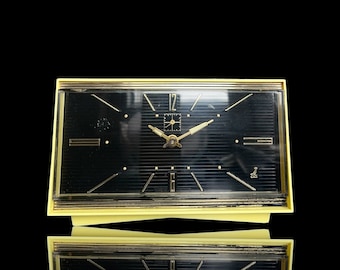 May include: A vintage rectangular clock with a black face and gold accents. The clock has a gold frame and gold hands. The clock is reflected in a black surface. The clock is likely from the mid-20th century.