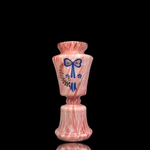 Puede incluir: Un jarrón de vidrio rosa y blanco con un lazo azul y un diseño floral. El jarrón tiene una base ancha y un cuello estrecho.