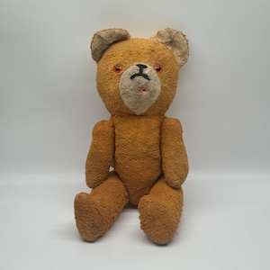 Può includere: Un orsacchiotto vintage con un esterno in pelliccia marrone usurata. L'orso ha occhi arancioni, un muso beige e un naso e una bocca cuciti in nero. Le sue braccia e gambe sono distese, dandogli un aspetto seduto.