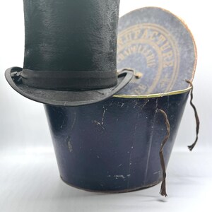 Peut inclure: Un haut-de-forme noir avec un ruban assorti posé sur une boîte à chapeau cylindrique bleu foncé. Le couvercle de la boîte est incliné, révélant le texte MANUFACTURE DE CHAPELLERIE imprimé en lettres dorées sur la surface circulaire bleue.