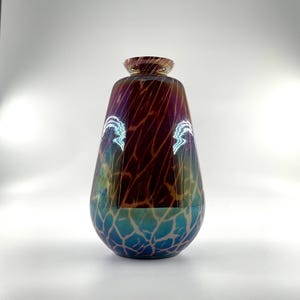 Puede incluir: Un jarrón decorativo de vidrio con un diseño único. El jarrón presenta una sección superior marrón oscuro con un patrón texturizado, que transita a una parte inferior azul y verde. El jarrón tiene una parte superior acampanada y una base redondeada.