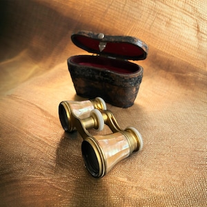 May include: Antique brass opera glasses with mother-of-pearl accents, closed and resting on a gold fabric surface. The glasses are in a brown leather case with a red velvet interior.