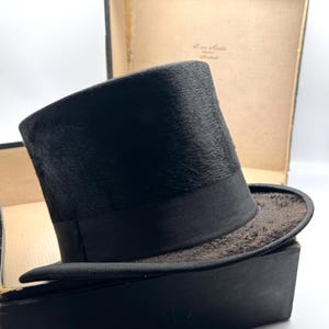 Peut inclure: Un chapeau haut de forme noir avec un ruban en gros-grain, posé dans sa boîte d'origine. Le chapeau a une surface légèrement texturée et un large bord. La boîte est sombre et usée.