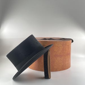 Peut inclure: Un chapeau haut de forme noir avec un ruban noir, une brosse à chapeau et une boîte à chapeau ronde et texturée. La boîte à chapeau est de couleur beige avec une bande marron foncé sur le dessus. La brosse a un manche en bois et des poils noirs.