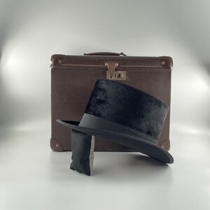 Peut inclure: Un chapeau haut de forme noir avec un ruban noir et une valise marron. Le chapeau est fait d'une matière douce et moelleuse. La valise est marron avec des protections d'angle en métal et un fermoir.
