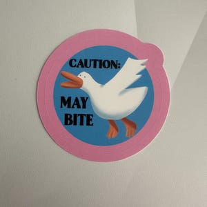 Puede incluir: Una pegatina redonda con borde rosa y centro azul que presenta un pelícano de dibujos animados. El pelícano es blanco con pico y patas naranjas. El texto "CAUTION: MAY BITE" está impreso en negro.