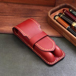 Könnte beinhalten: Ein rotes Leder-Stifteetui mit Klappenverschluss und Ziernähten. Das Etui ist für zwei Stifte konzipiert. Der Hintergrund umfasst ein Holztablett mit Stiften und eine Holzoberfläche.