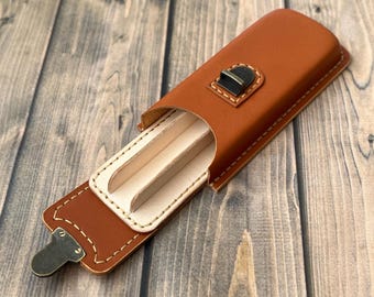 May include: A brown leather case with a flap that opens to reveal two slots for holding items. The case has a brass clasp and stitching around the edges.