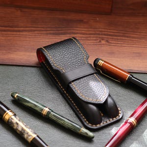 Könnte beinhalten: Ein schwarzes Leder-Stiftetui mit gelben Nähten, das zwei Stifte enthält. Drei zusätzliche Stifte in Schwarz, Grün und Rot mit goldenen Akzenten sind um das Etui herum angeordnet. Die Stifte liegen auf einer dunkelgrünen Oberfläche.