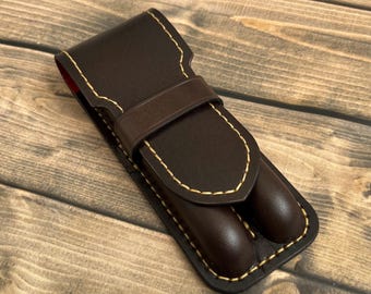 May include: A brown leather pen case with a flap closure and a stitched design. The case has two slots for pens.