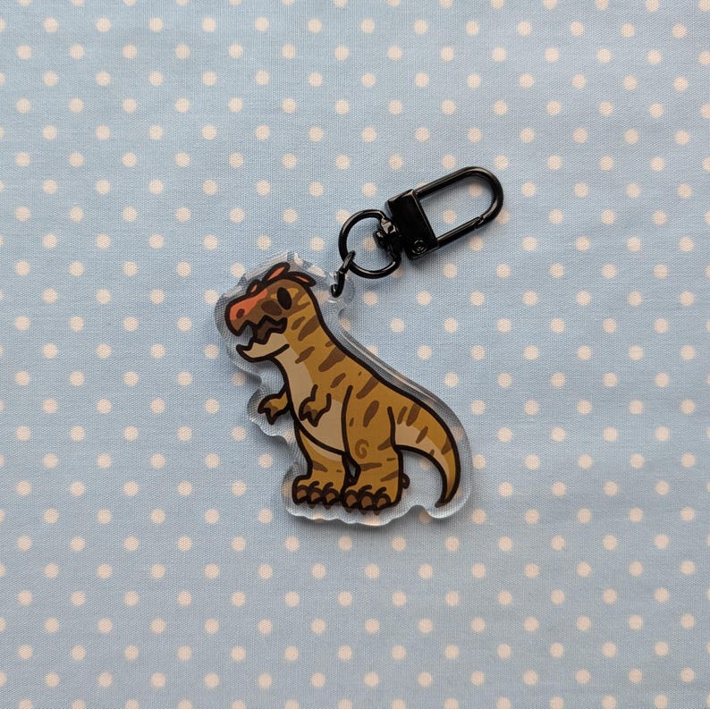 May include: A clear acrylic keychain featuring a cartoon dinosaur with brown and tan stripes. The dinosaur has a red crest and a black eye. The keychain is attached to a black metal ring.