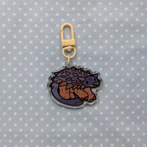 May include: A light blue and brown cartoon Ankylosaurus keychain with a white metal key ring. The keychain is made of clear acrylic.