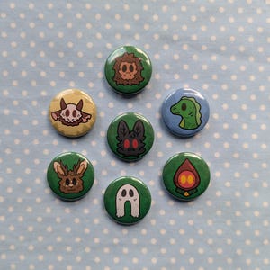 May include: Seven round pin badges featuring quirky illustrations. Designs include a lion-like creature, a skull, a moth, a dinosaur, a rabbit, a ghost, and a figure with a flame-like head. The badges are set against a light blue background with white polka dots.