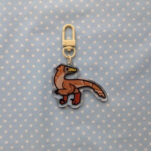 May include: A brown and white dinosaur keychain with a yellow beak and red feet. The keychain is attached to a gold metal ring.