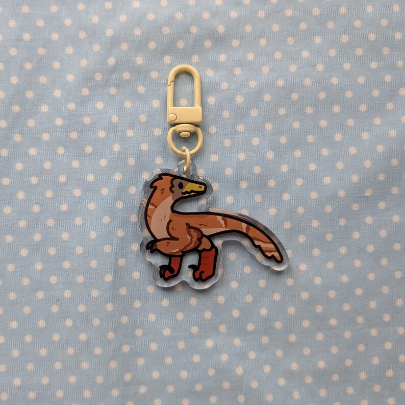 May include: A brown and white dinosaur keychain with a yellow beak and red feet. The keychain is attached to a gold metal ring.
