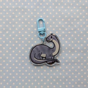 May include: A blue and gray dinosaur keychain with a light blue metal clasp. The dinosaur is standing on all fours and has a long neck.
