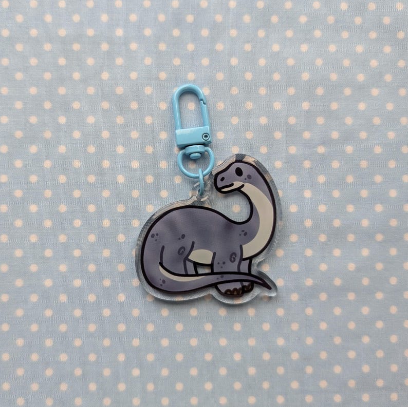 May include: A blue and gray dinosaur keychain with a light blue metal clasp. The dinosaur is standing on all fours and has a long neck.